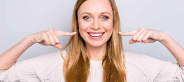 Portrait of joyful satisfied girl gesturing her beaming white healthy teeth with two forefingers looking at camera isolated on grey background. Orthodontic concept